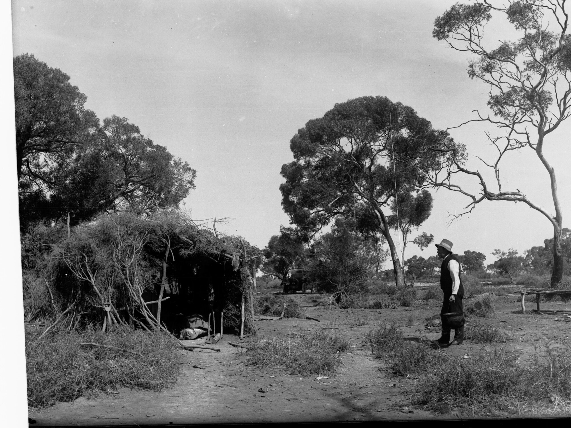 Rural view showing hut made of branches, man holding bucket, also showing is automobile