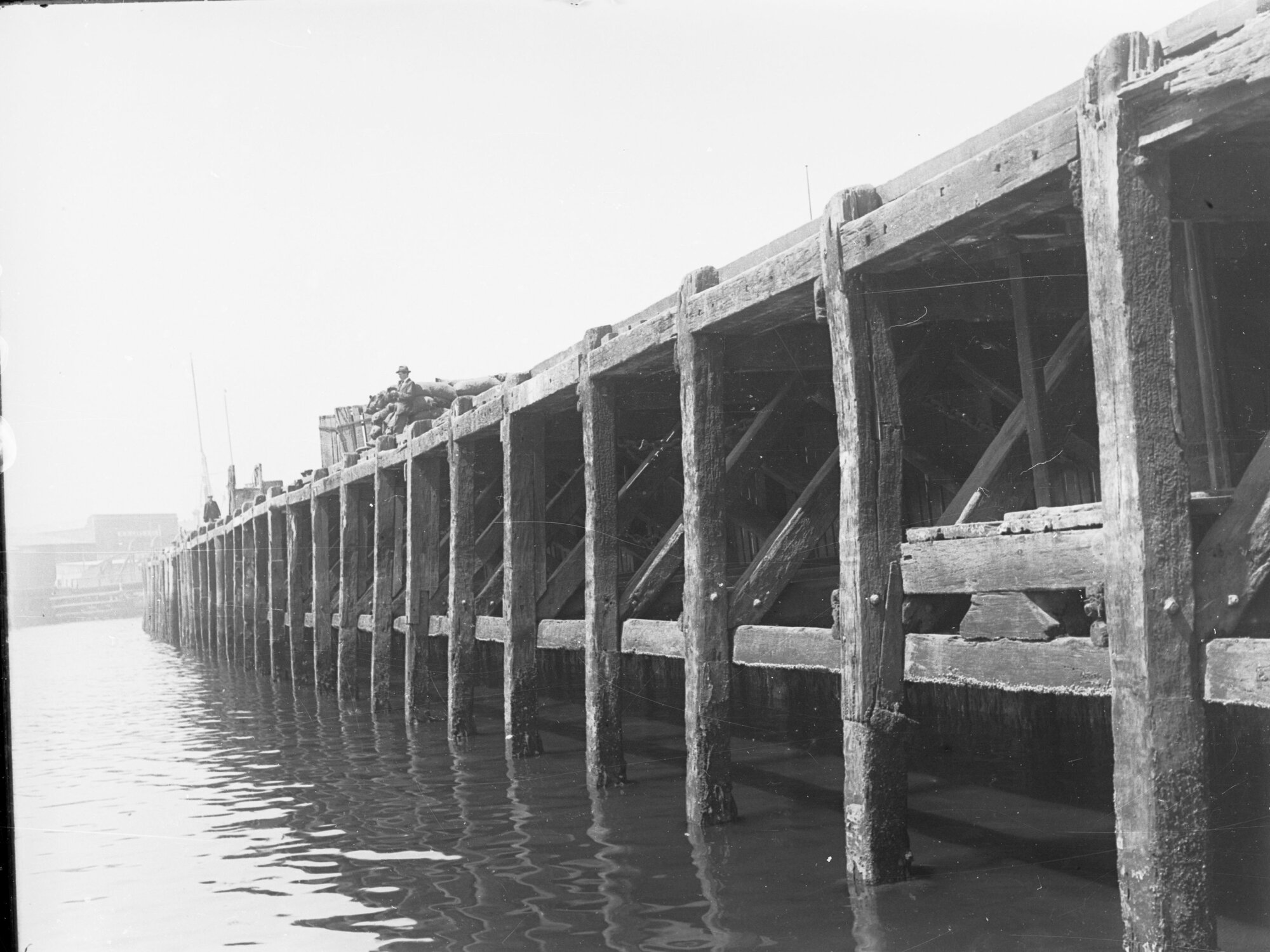 No. 2 Quay at Port Adelaide - view of underside of wharf - building in background W R Cave &amp; Son, and man fishing