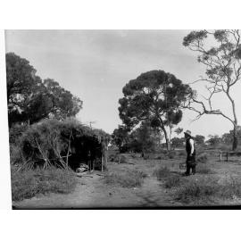 Rural view showing hut made of branches, man holding bucket, also showing is automobile